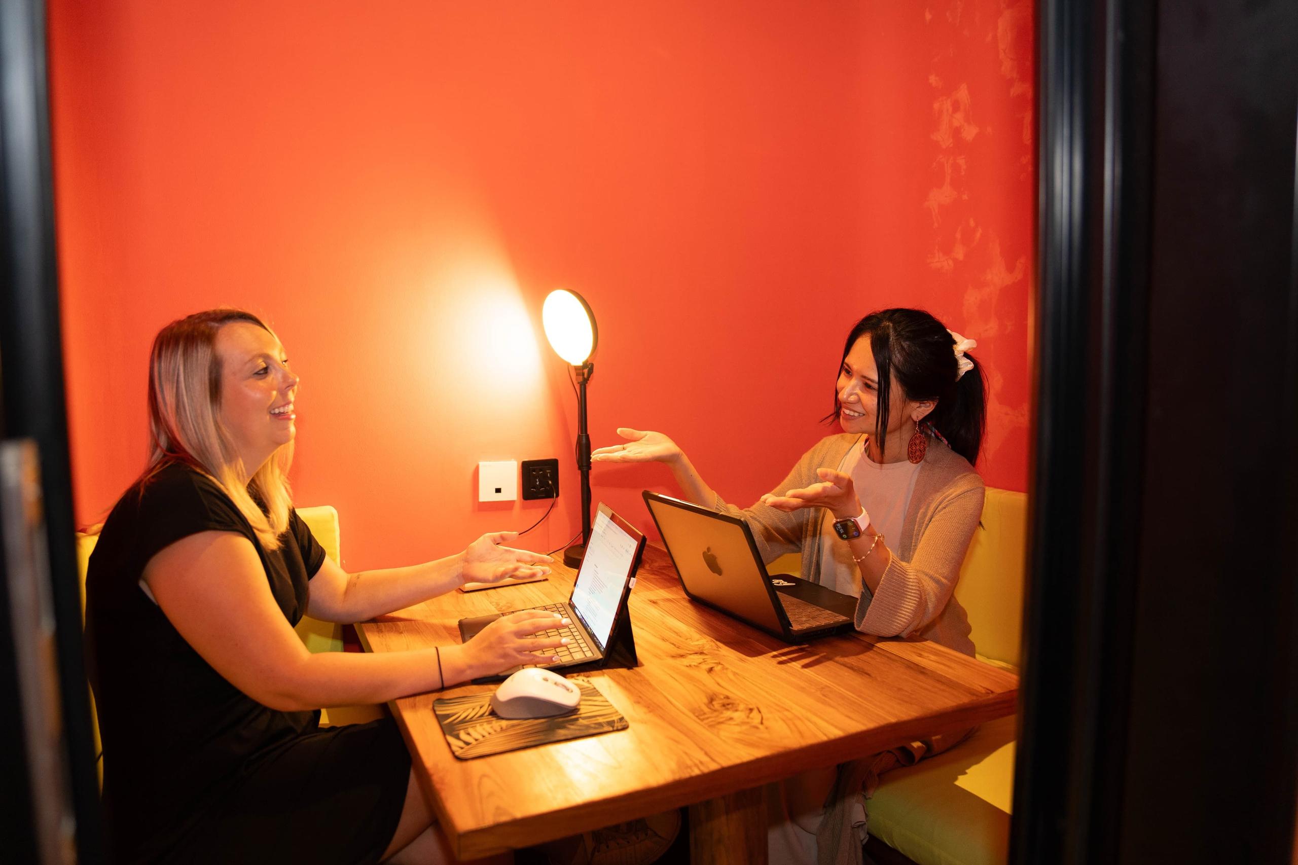 Two women working on laptops in a small focus room with orange walls and a ring light