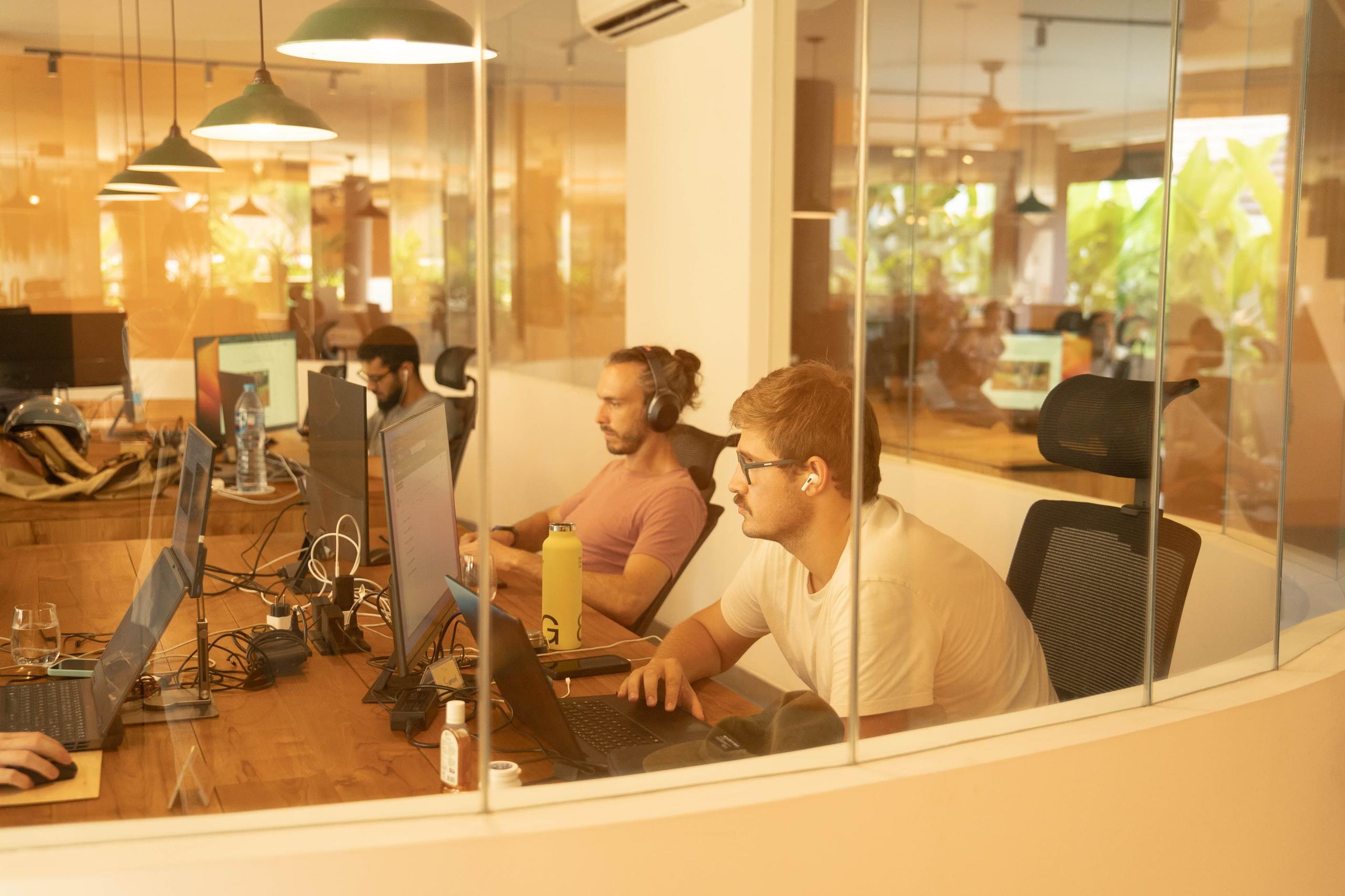 Members working at desks in the indoor coworking area, viewed through glass partition