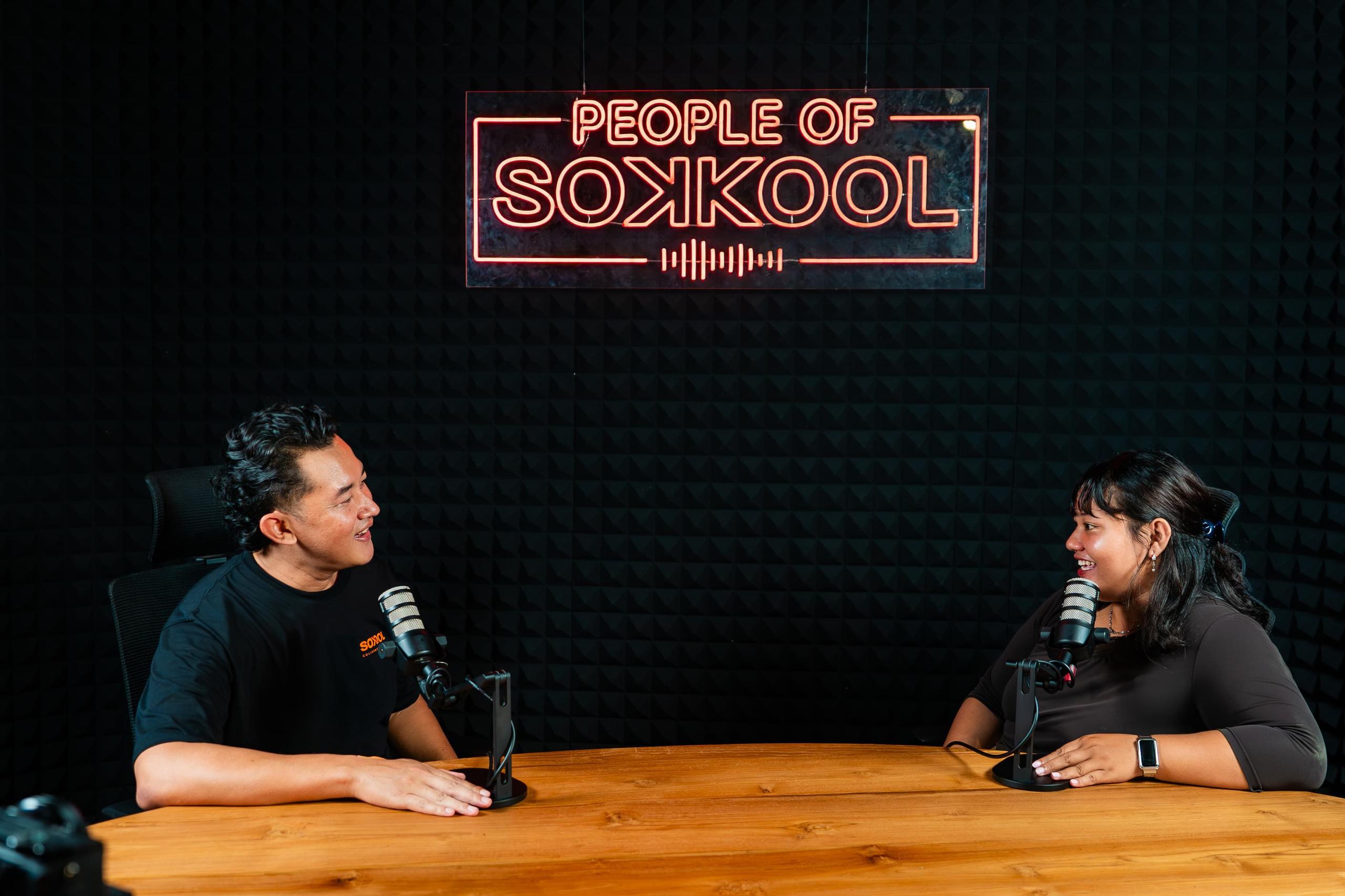 Two people recording a podcast in the studio with 'People of Sokkool' neon sign in the background