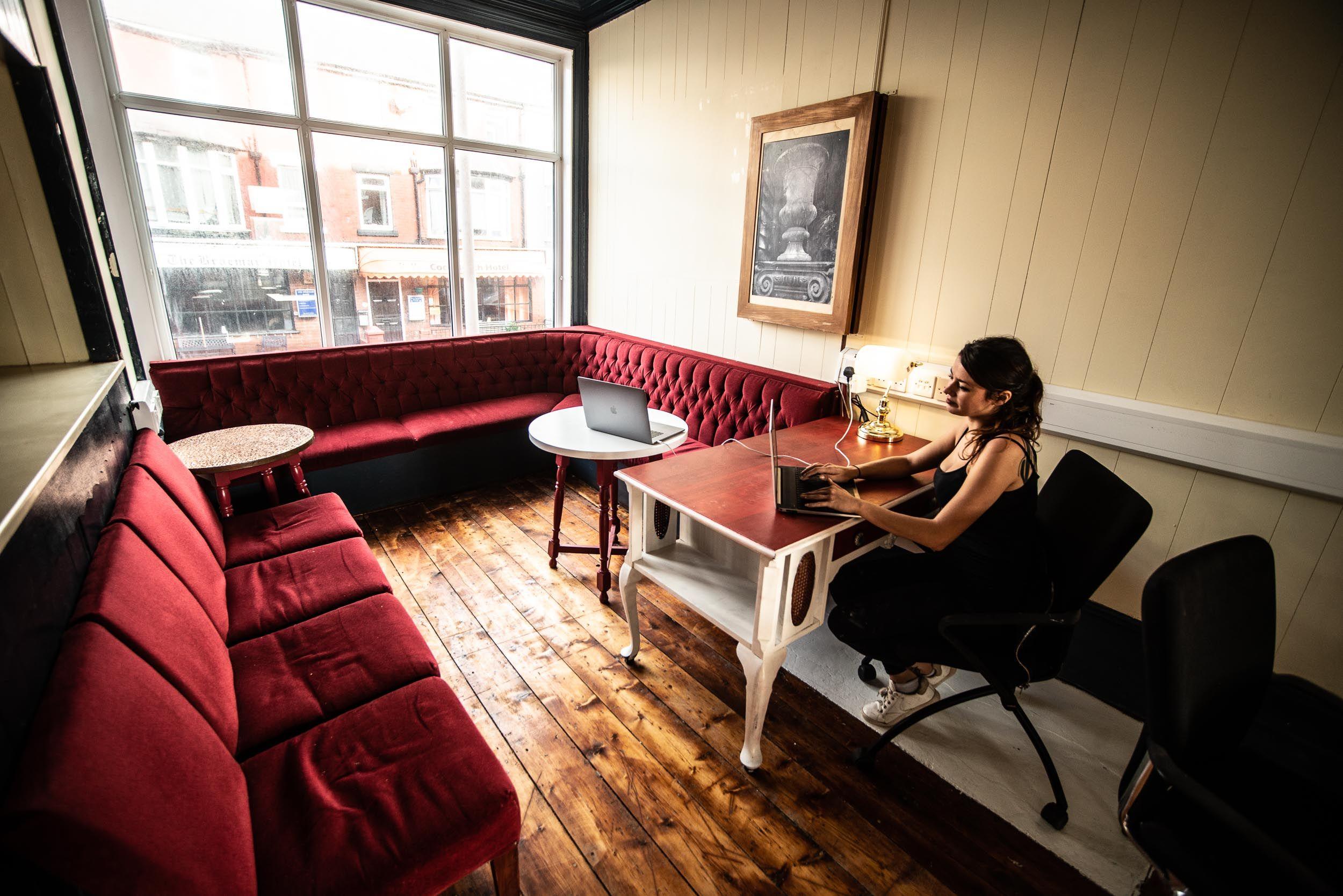 Person working at a desk in the main coworking room with red sofa bar area and large bay window