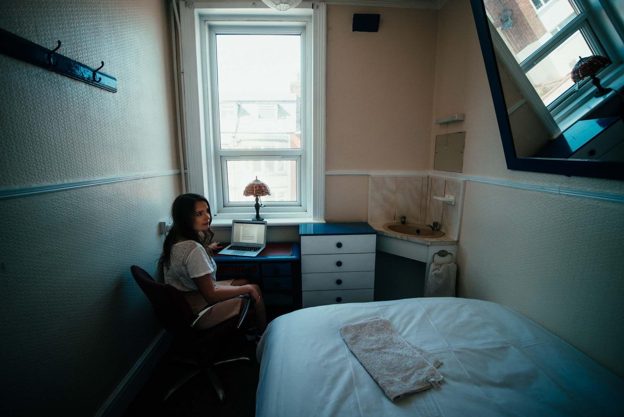 Person sitting at a small desk with a laptop in a single bedroom with single bed