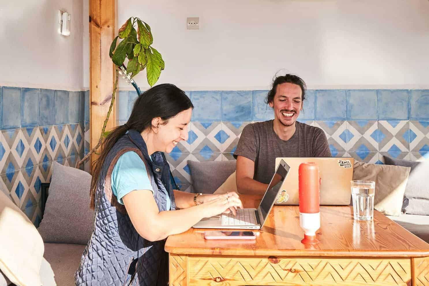Two coworkers laughing and working on laptops in the SunDesk living area with blue Moroccan tile decor