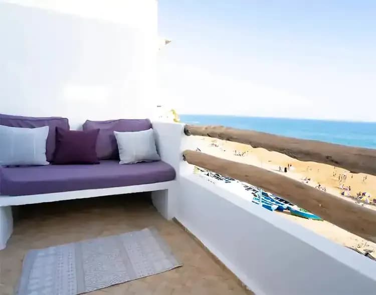 Cozy balcony with a purple couch and cushions overlooking a sandy beach and blue ocean under a clear sky at SunDesk Taghazout