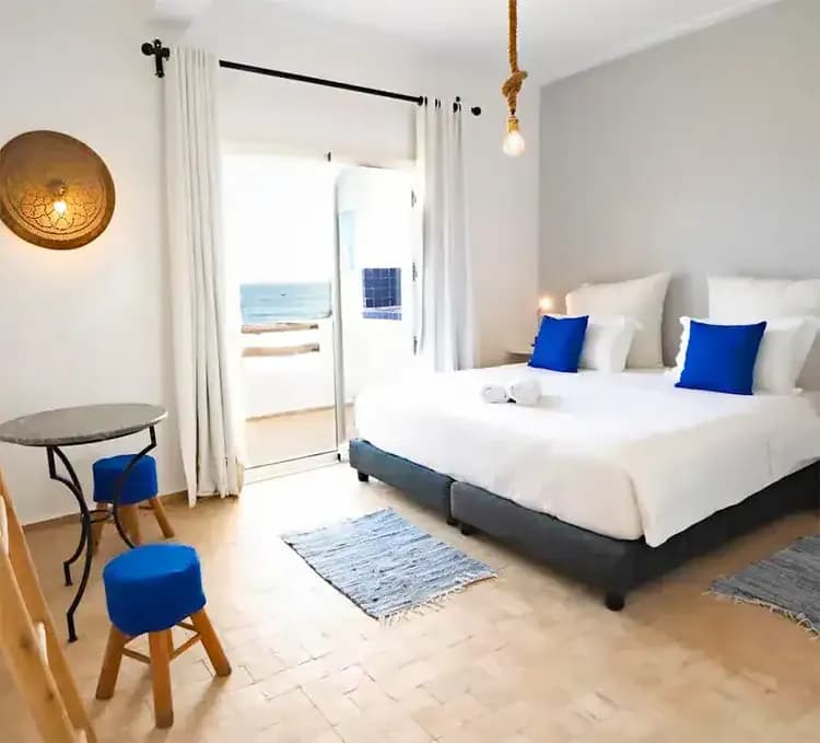 Bright, modern bedroom with a double bed, blue accents, minimalist decor, a table, and a balcony view of the ocean in SunDesk Taghazout