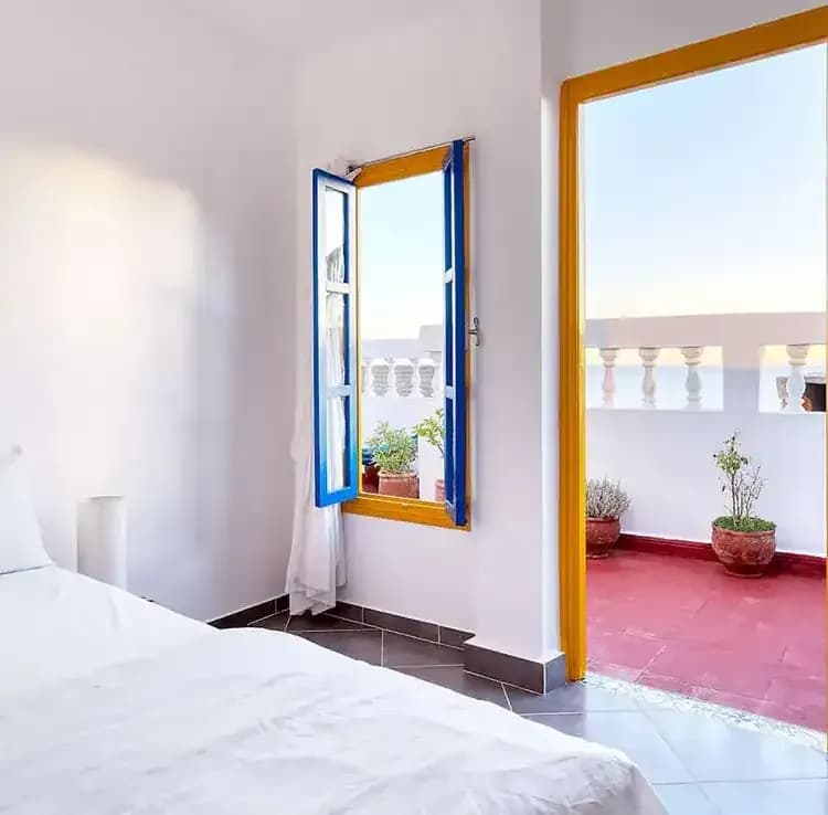 Bright bedroom with a white bed, blue window frames, and a doorway leading to a sunny terrace with plants at SunDesk in Taghazout