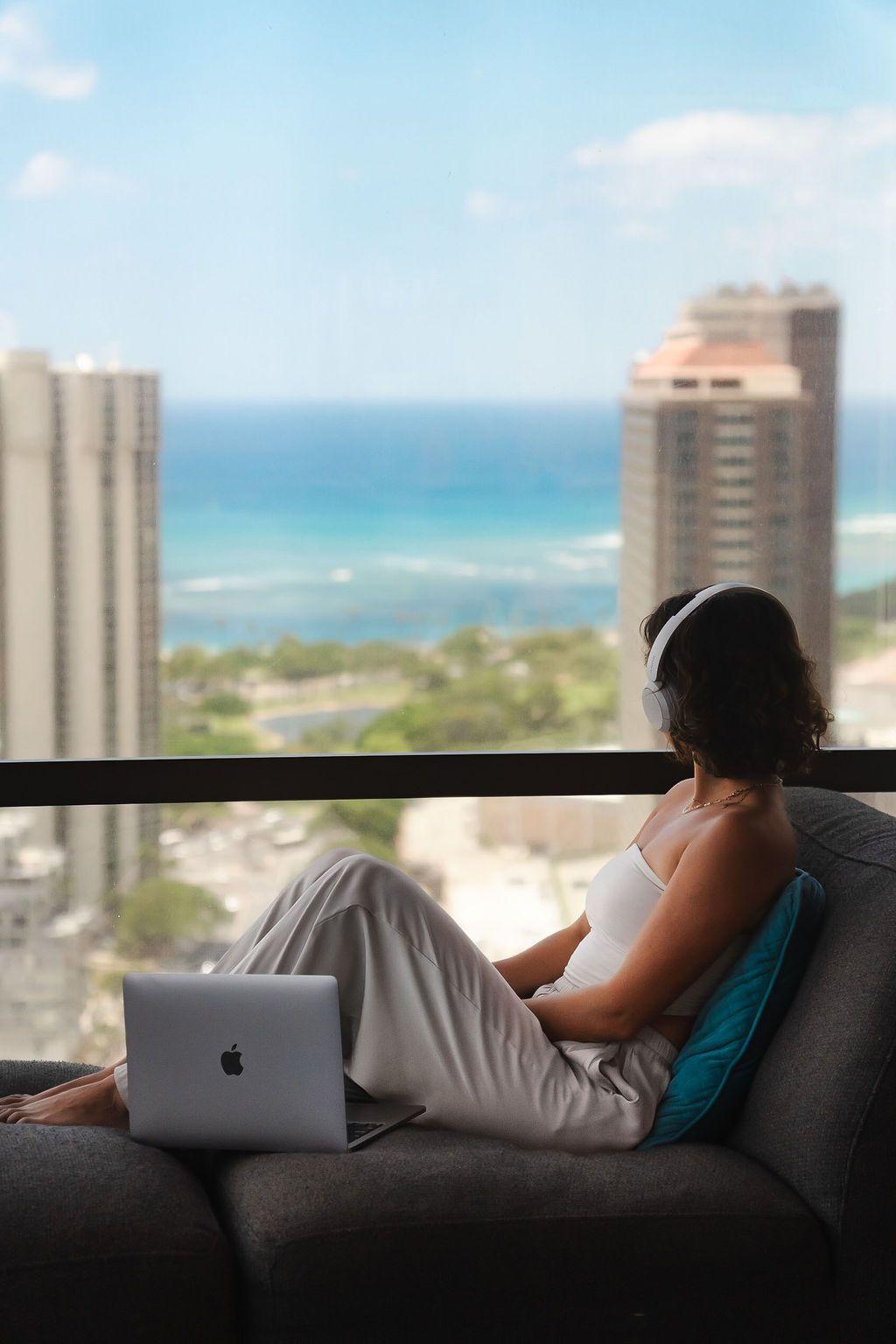Member with headphones working on a laptop on the lounge sofa with panoramic ocean and city views through floor-to-ceiling windows