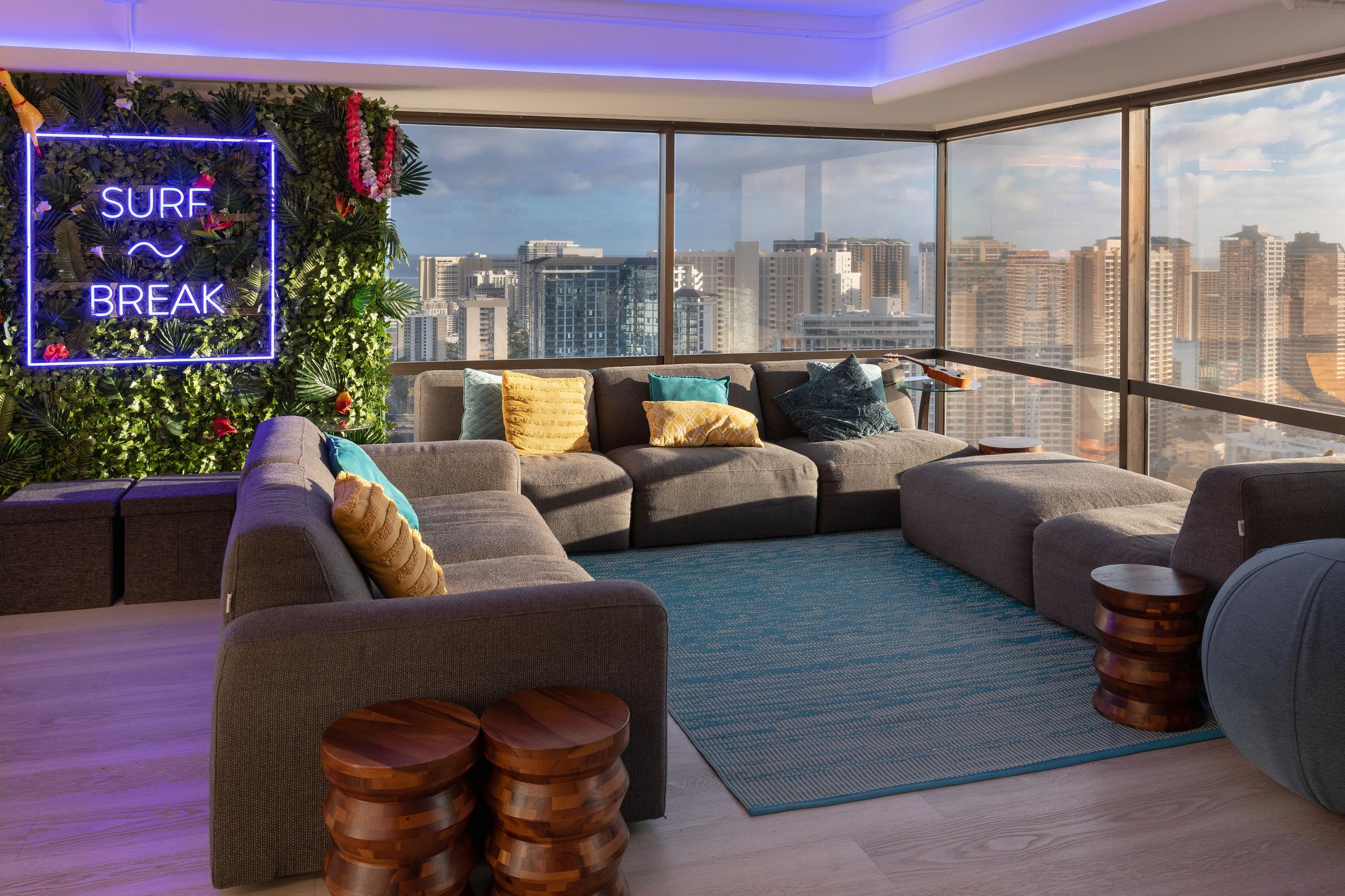 Spacious lounge area on the 40th floor with large sectional sofa, Surfbreak neon sign on a living plant wall, and floor-to-ceiling windows overlooking Honolulu