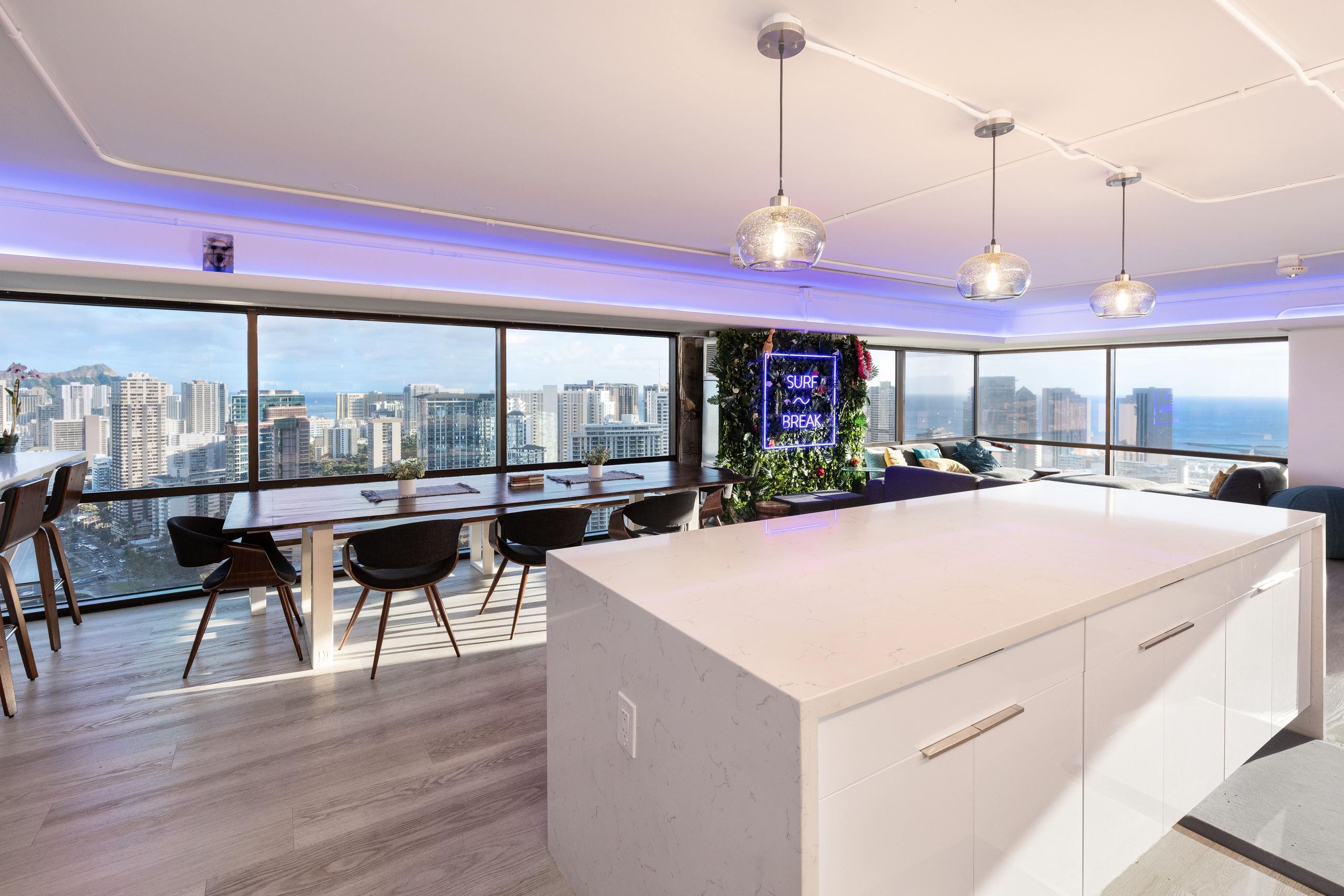 Fully-equipped shared kitchen with large white island and open-plan dining area with floor-to-ceiling windows overlooking Honolulu skyline