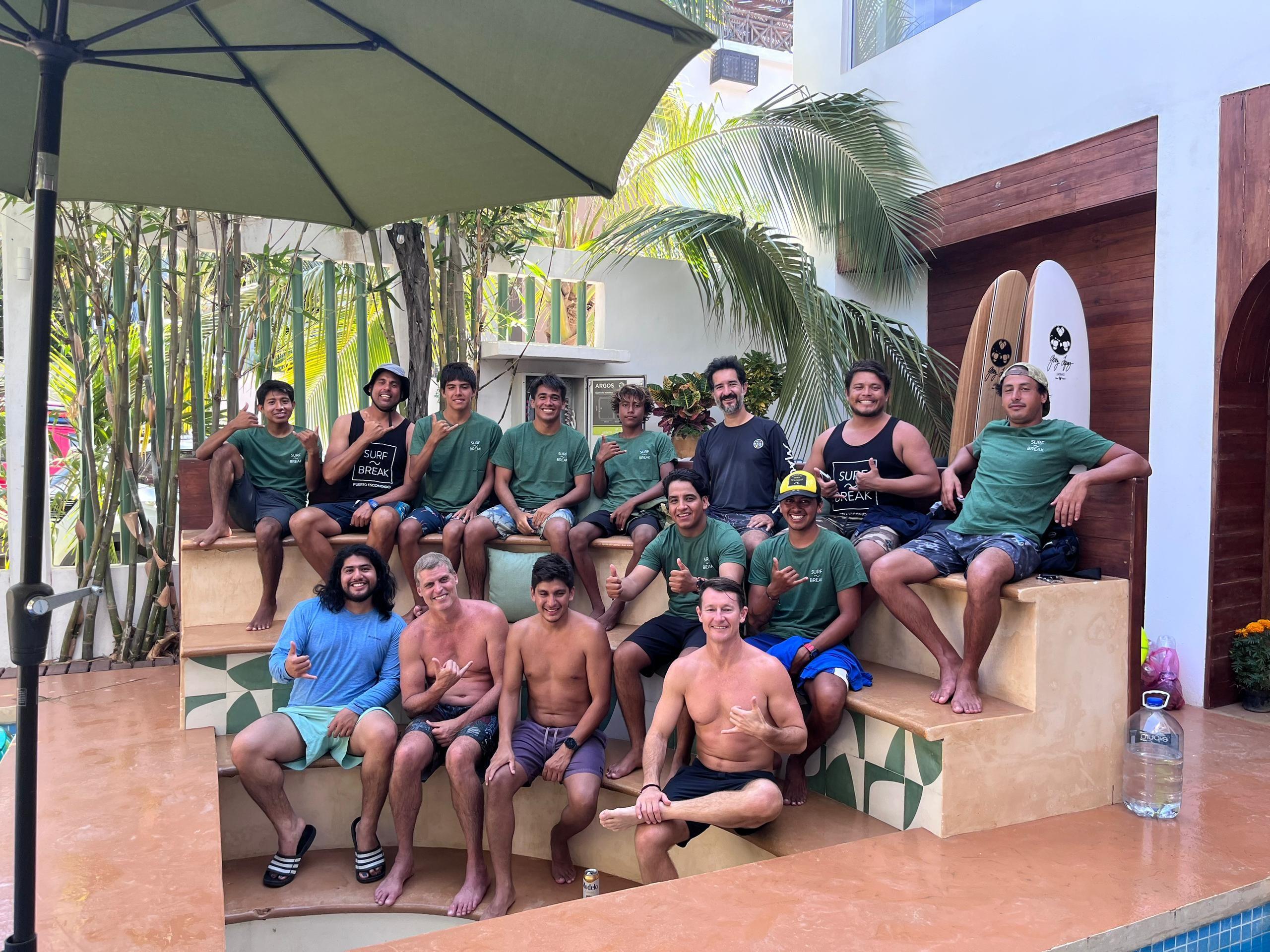 Large group of people posing together by the pool and sunbathing area at Surfbreak PXM, with surfboards visible
