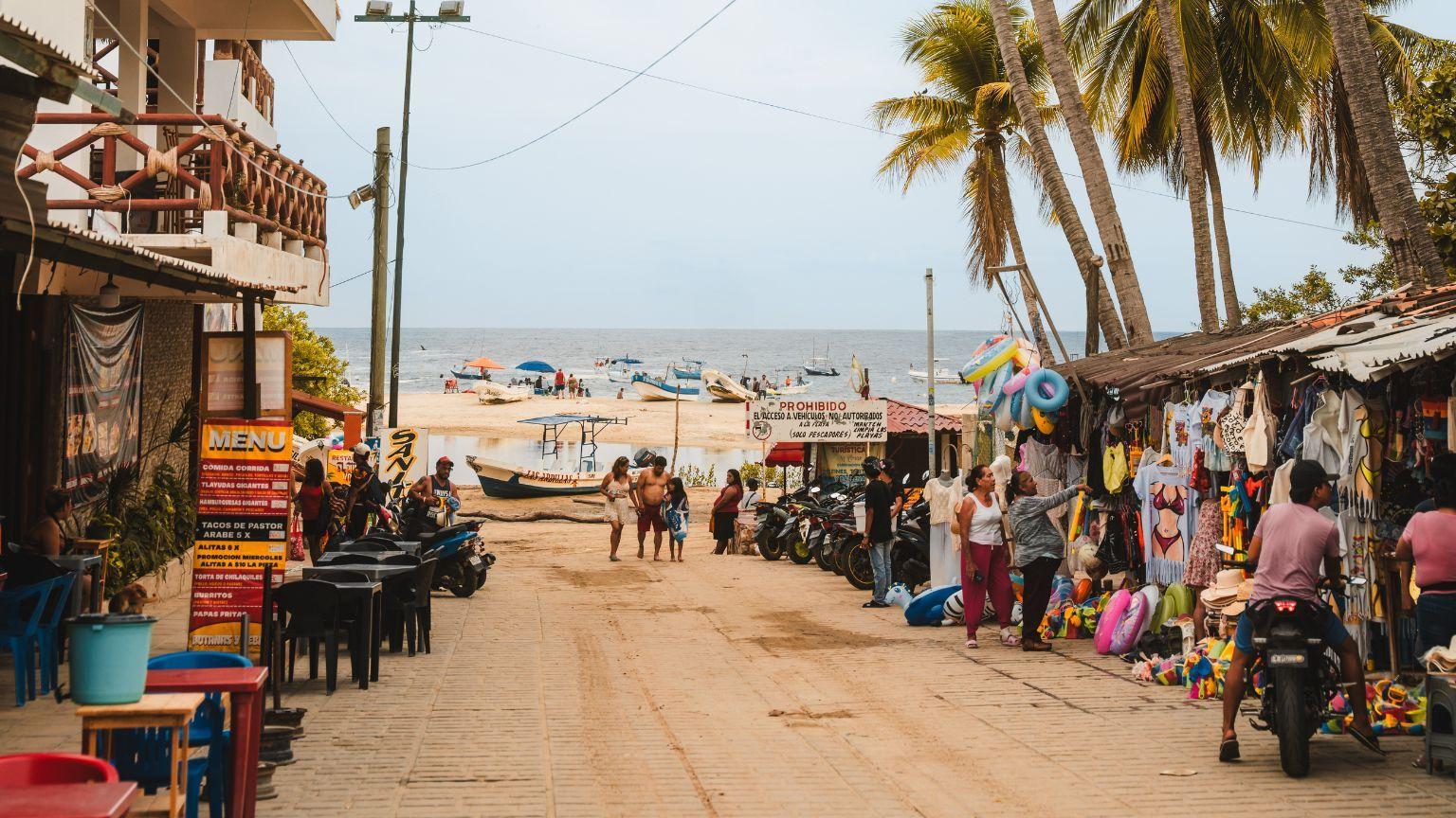 A street-level view of a local market in Puerto Escondido with shops, palm trees, and the beach in the background