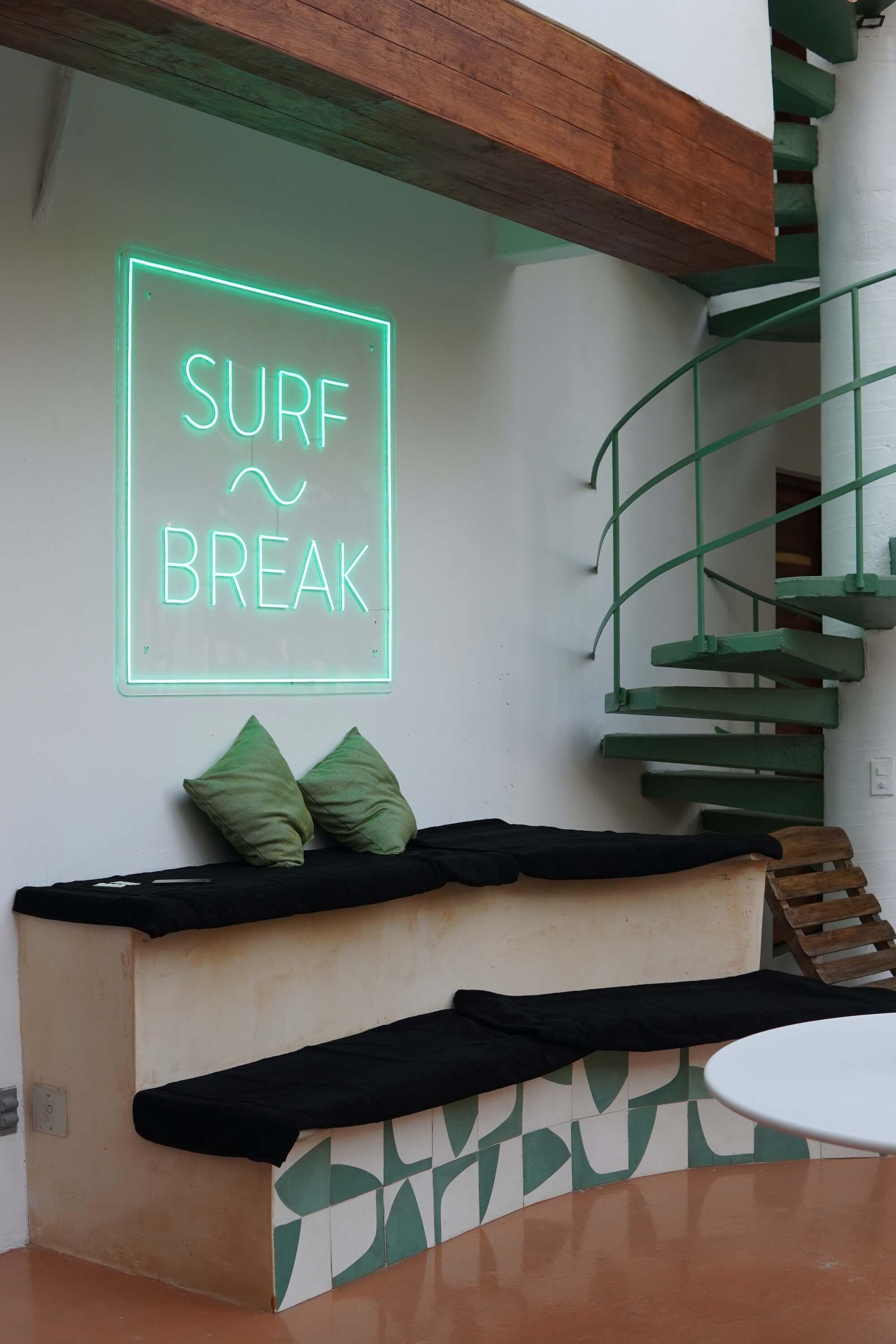 Interior social lounge area with a glowing 'Surf Break' neon sign, bench seating with green cushions, and a spiral staircase