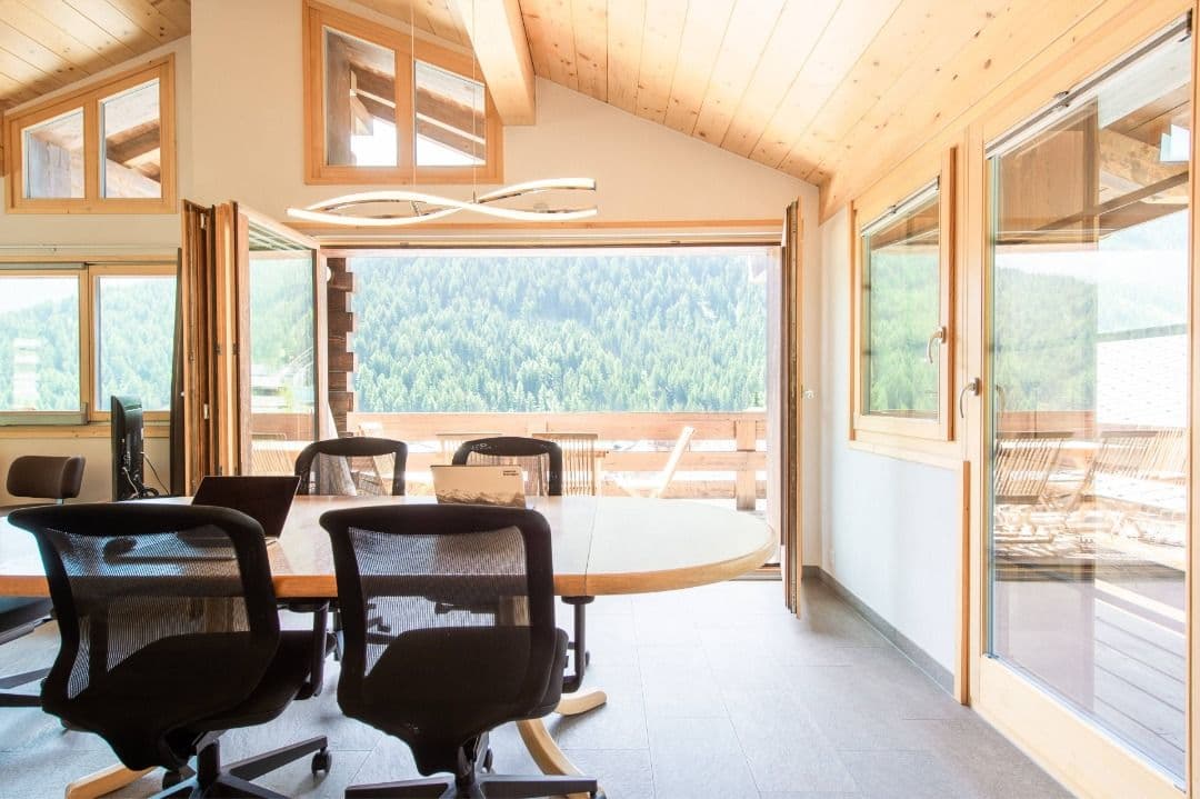 Coworking area in the chalet with ergonomic chairs, large table, and open folding doors leading to the terrace with forest views