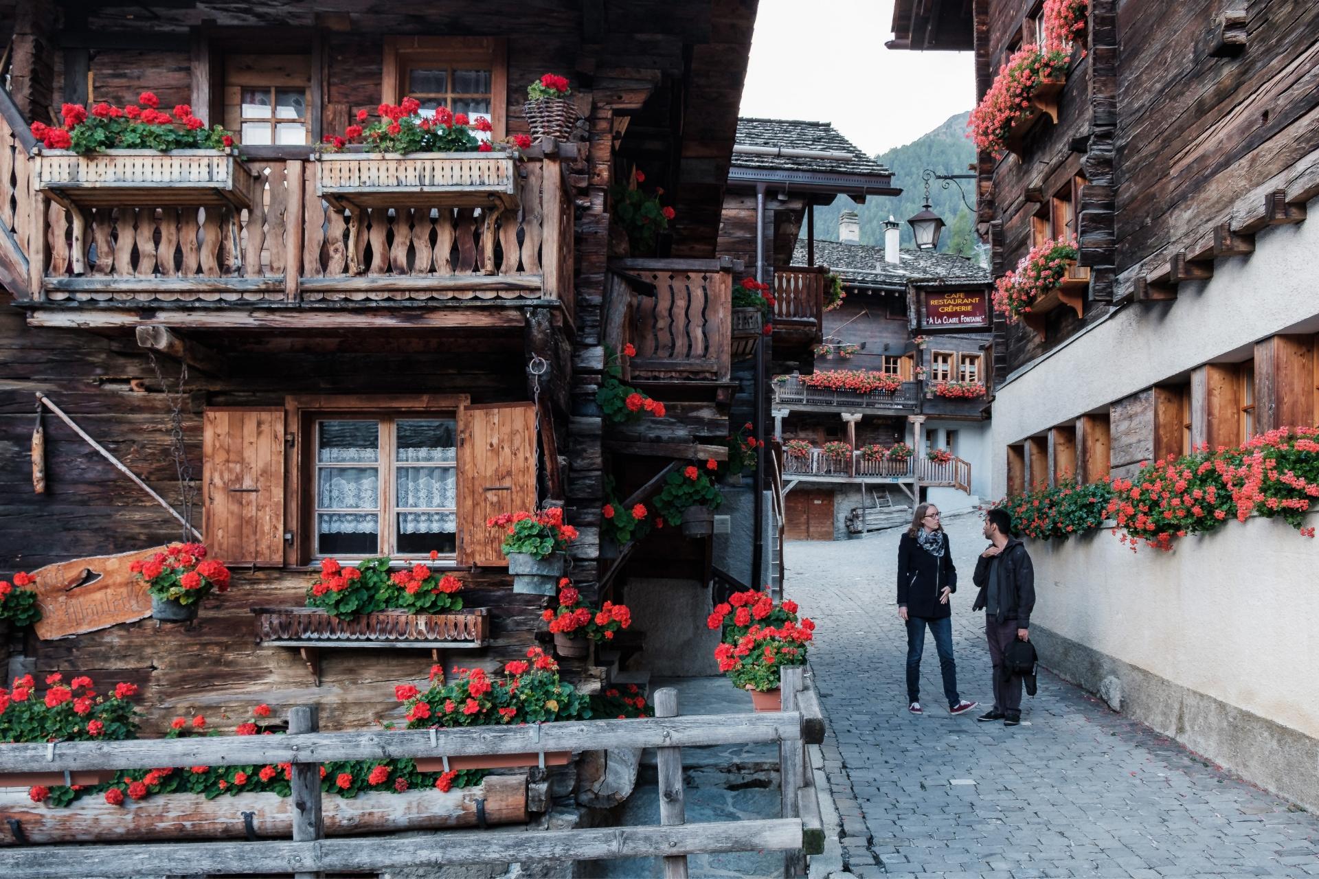 Two people walking in the old village streets of Grimentz with traditional Swiss chalets decorated with red geraniums
