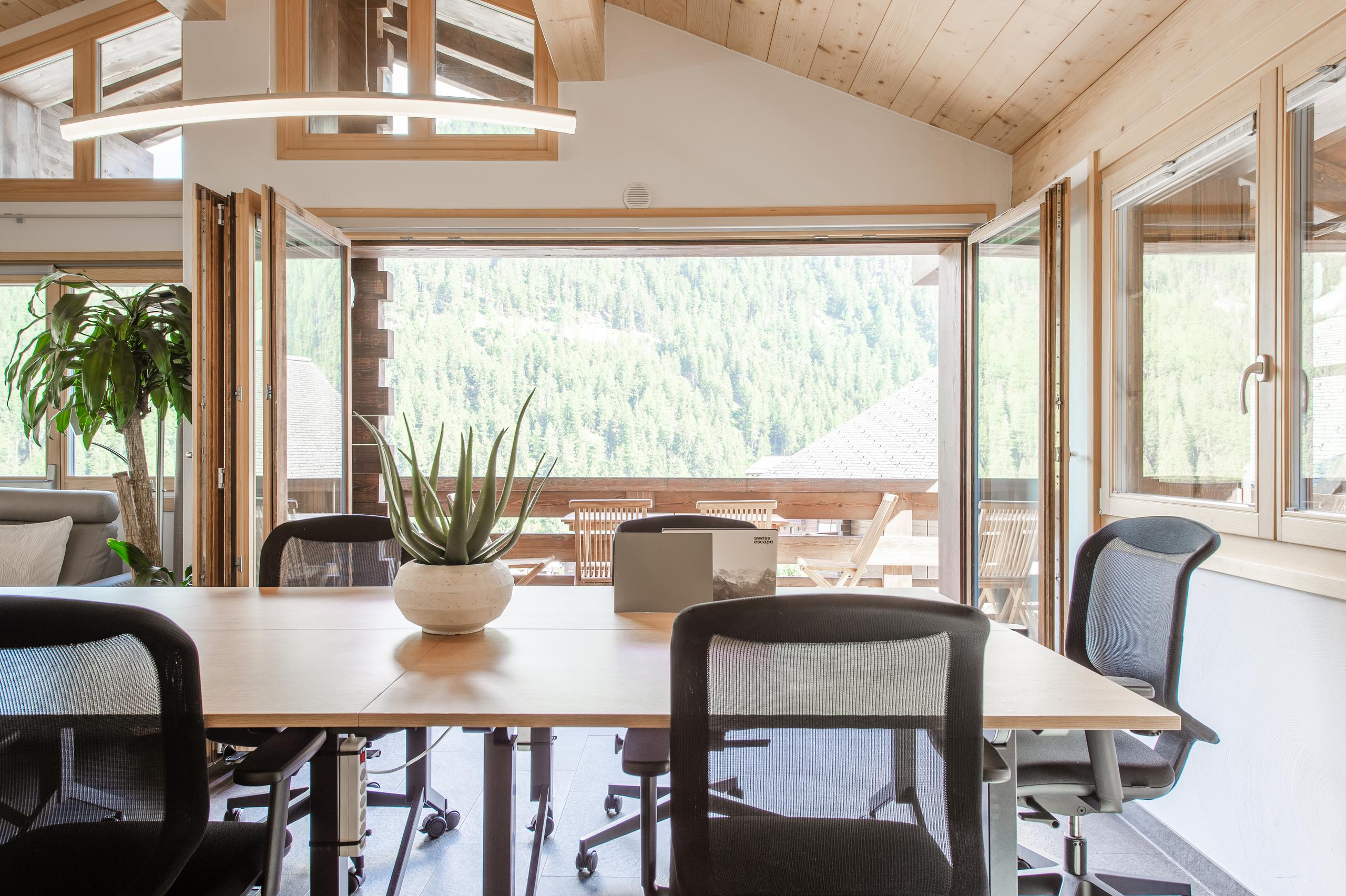 Communal coworking area in the Swiss Escape chalet with ergonomic chairs, large desk, and panoramic mountain views through open windows