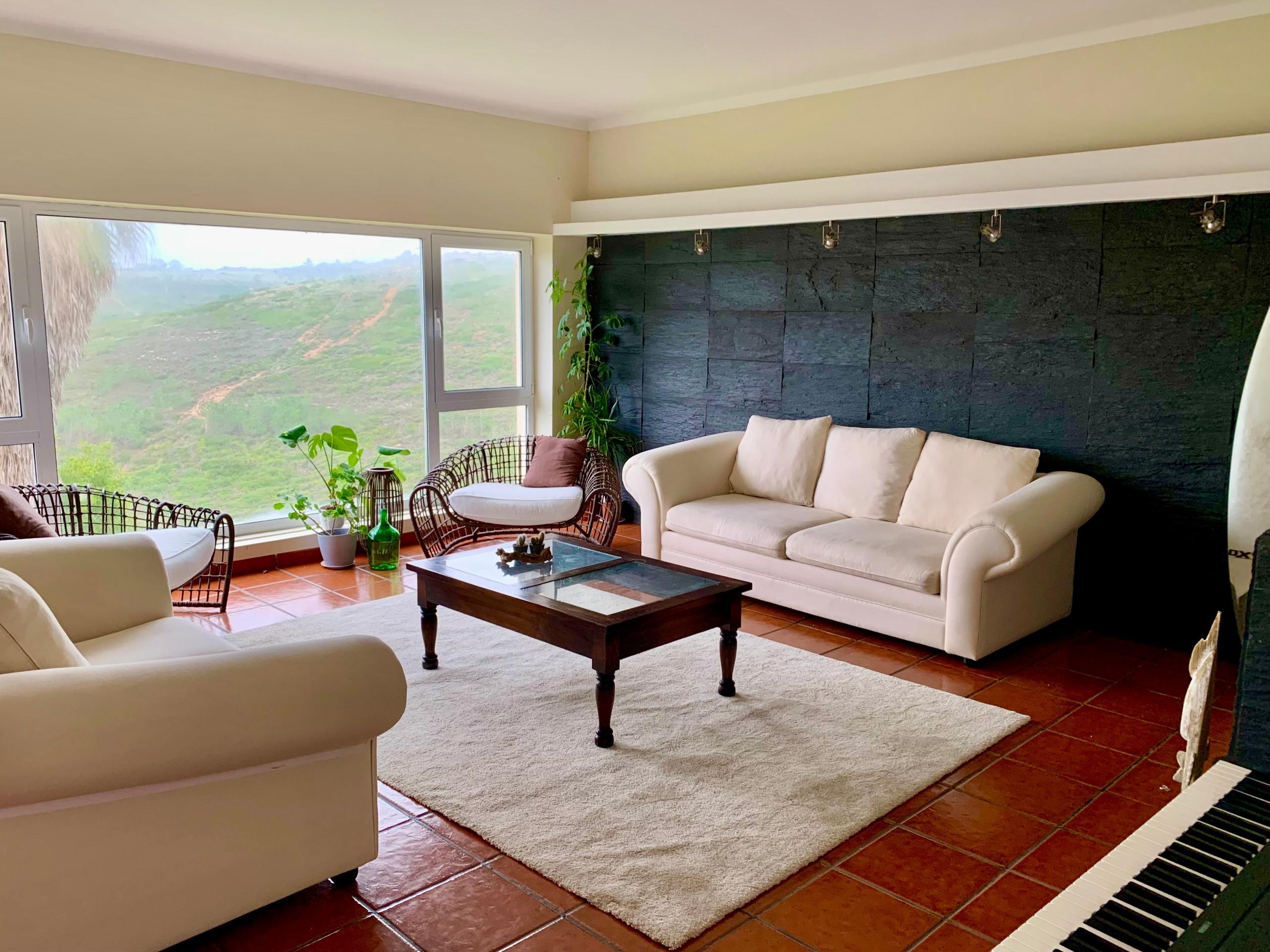 Spacious living room with large white sofas, a coffee table, and large windows overlooking green hills