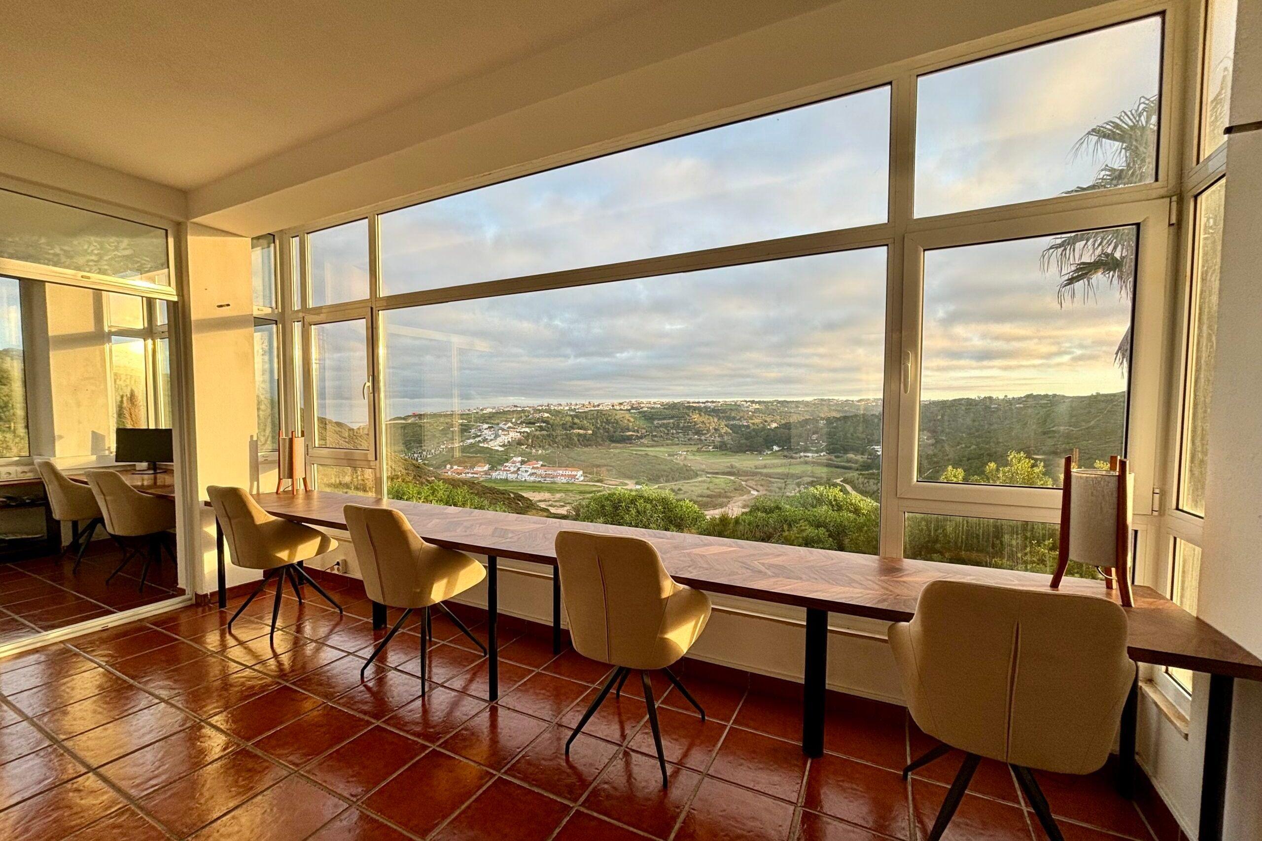 Coworking space with long bench-style desks and chairs facing large floor-to-ceiling windows with panoramic ocean and valley views