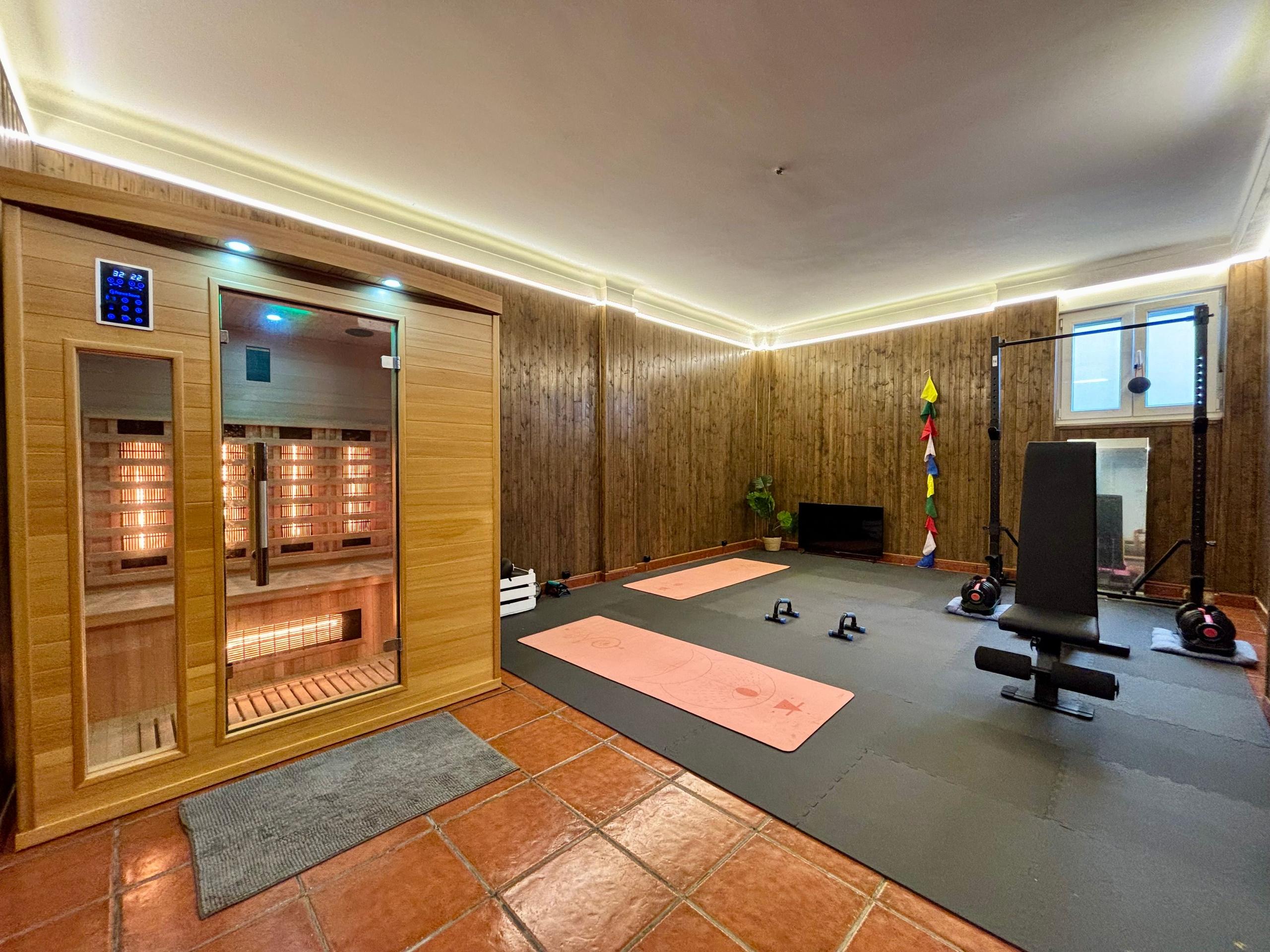 Combined gym and infrared sauna space with yoga mats, power rack, bench, and sauna cabin with wood-panelled walls