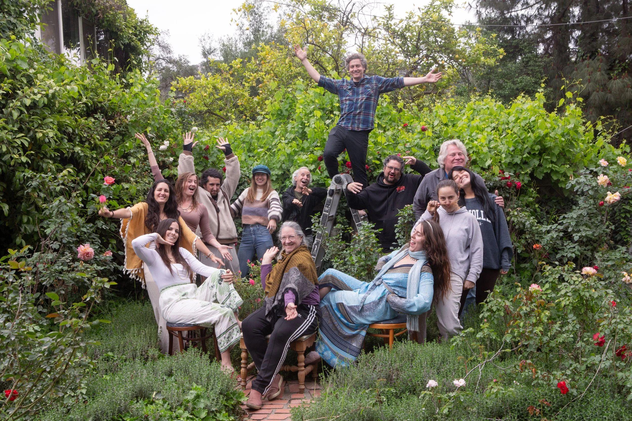 A diverse group of people posing in a garden with trees and flowering bushes, an older man on a ladder at center
