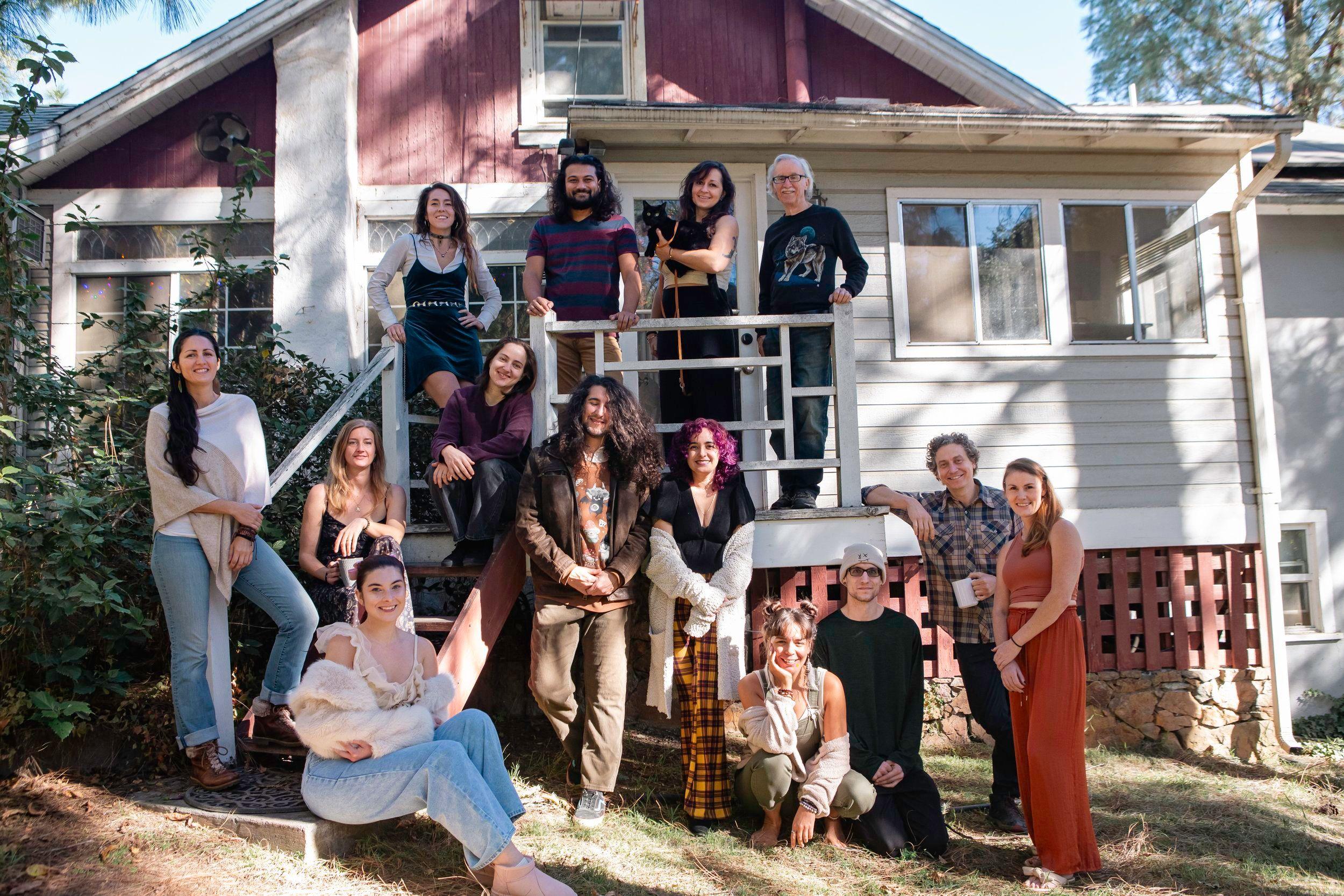 A group of people in front of a house, some sitting on ground, others on stairs, with a black cat being held. House has red upper part, white lower part, multiple windows