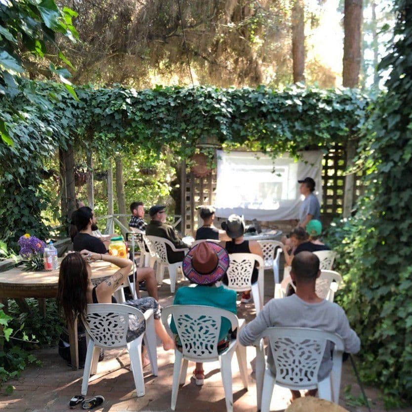 People sitting outdoors on chairs watching a presentation or movie set up under a leafy canopy in a garden setting
