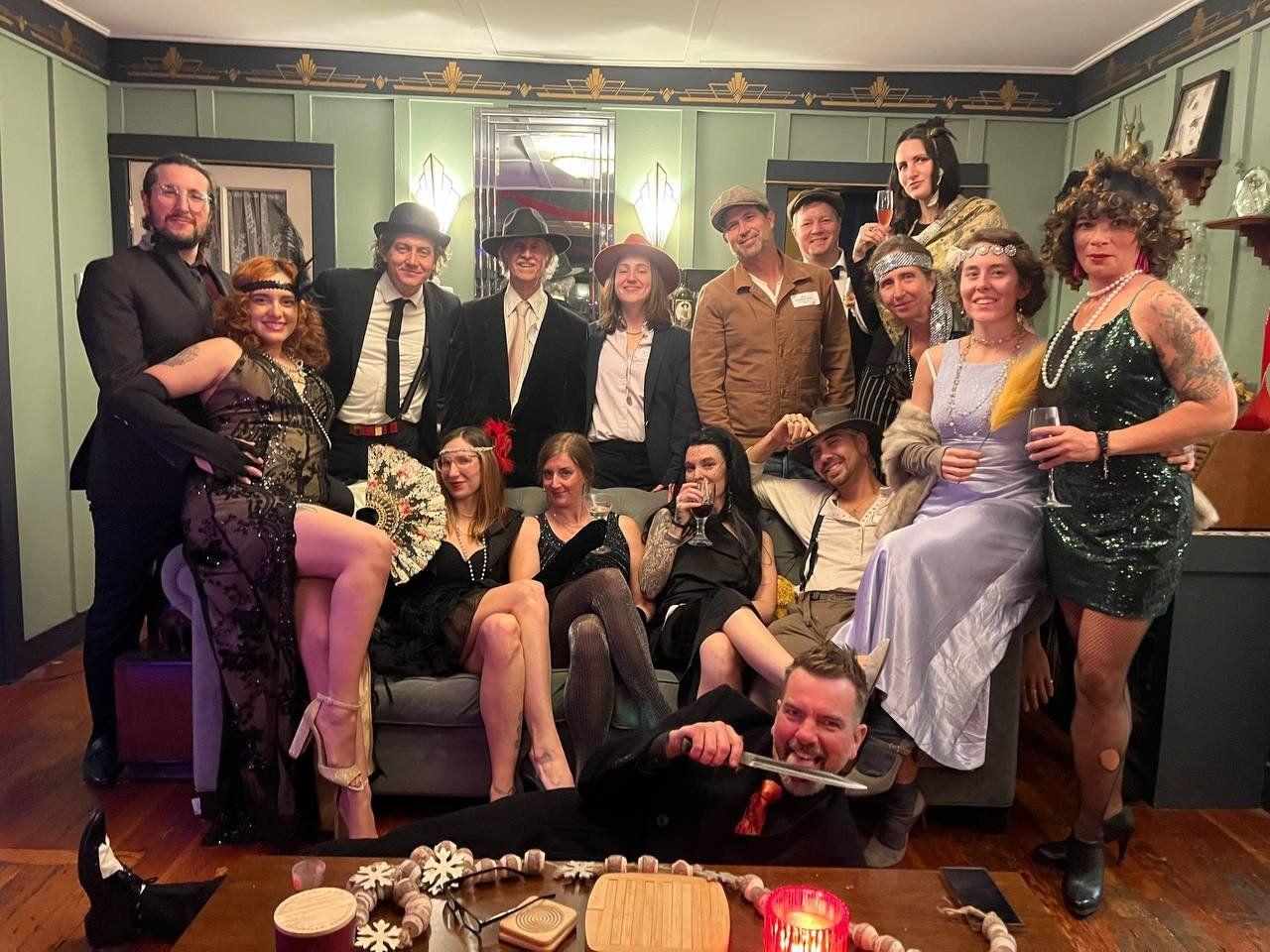 A group of people dressed in vintage and flapper-era costumes gathered in a living room, celebrating with drinks during a themed party