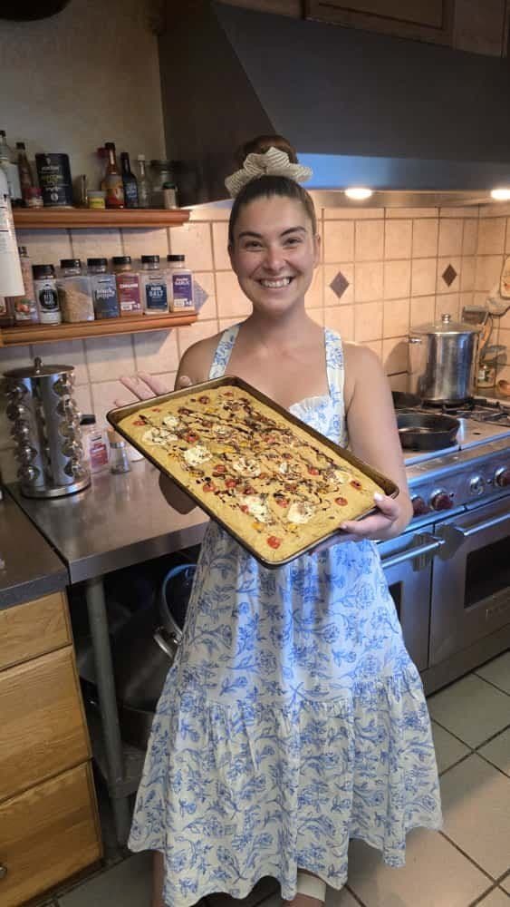 A woman in a blue and white floral dress holding a baking tray of a decorated dessert in a kitchen