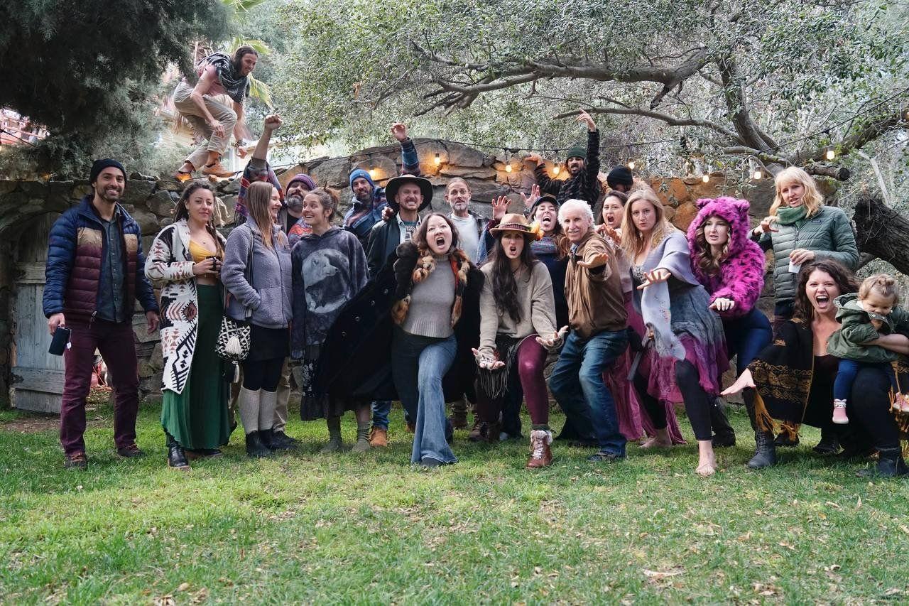 A group of people gathered outdoors on a grassy area, some making playful poses, with a large tree and string lights overhead